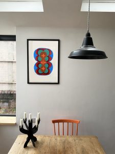 View of a room with the print displayed in a black frame on a wall. There is a black lamp shade hanging to the right of the frame and an orange chair that matches the orange in the print.